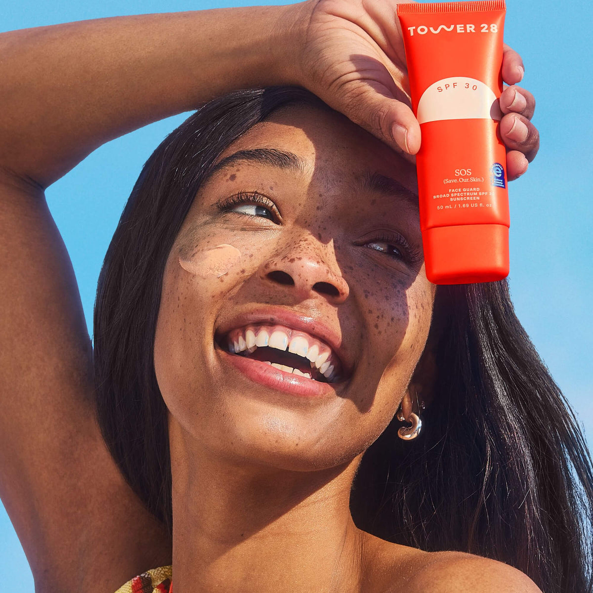 A person smiles while holding up a bottle of Tower 28 Beauty's SOS FaceGuard SPF 30. A swatch of the product is visible on their cheek.  [Shared: SOS FaceGuard SPF 30] 