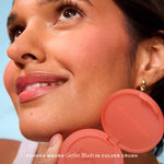 A person holds Tower 28 Beauty’s GetSet Powder Blush in the shade Culver Crush with a swatch of pinky peach on their cheek. [Culver Crush]