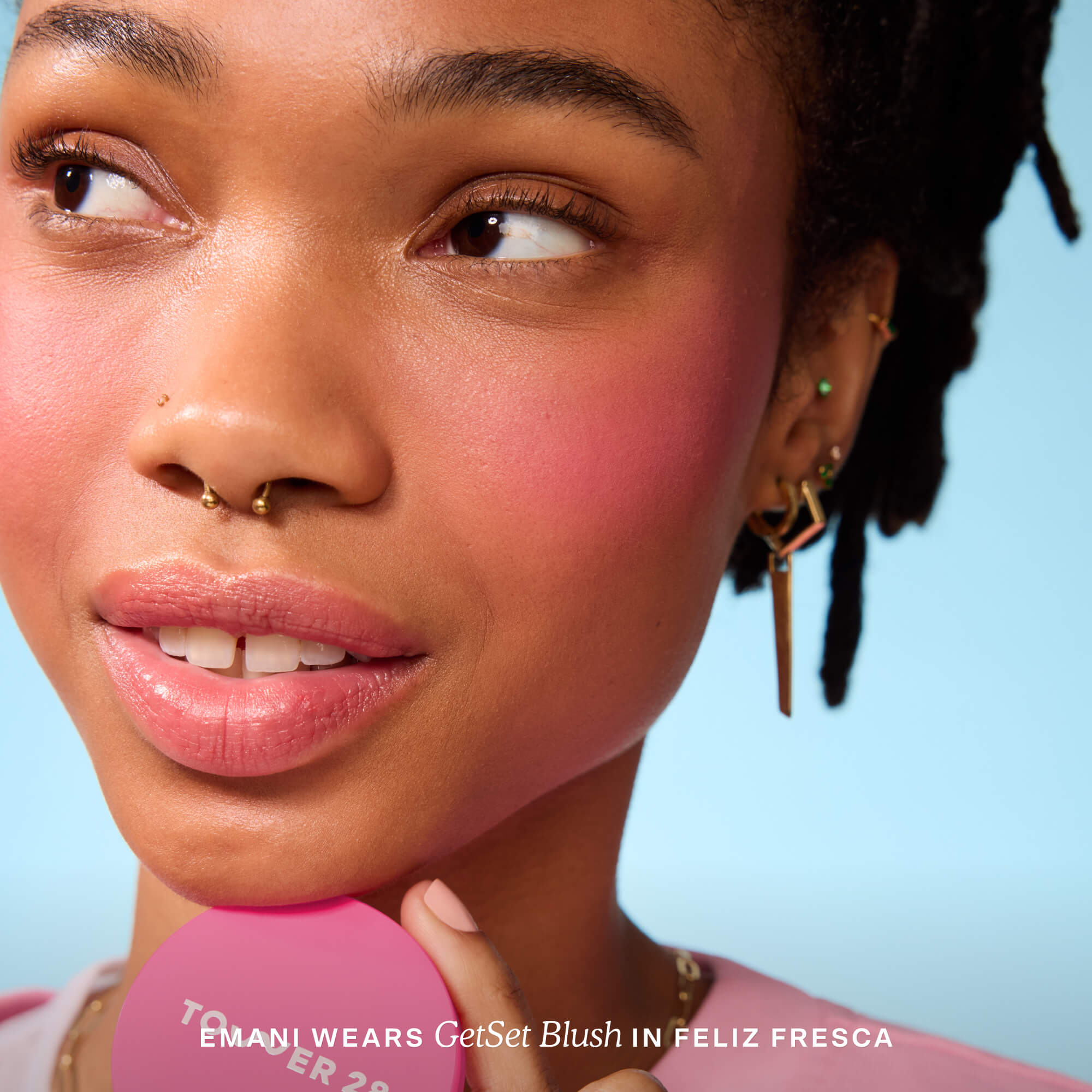 A person holds Tower 28 Beauty’s GetSet Powder Blush in the shade Feliz Fresca with a swatch of warm hot pink on their cheek. [Feliz Fresca]