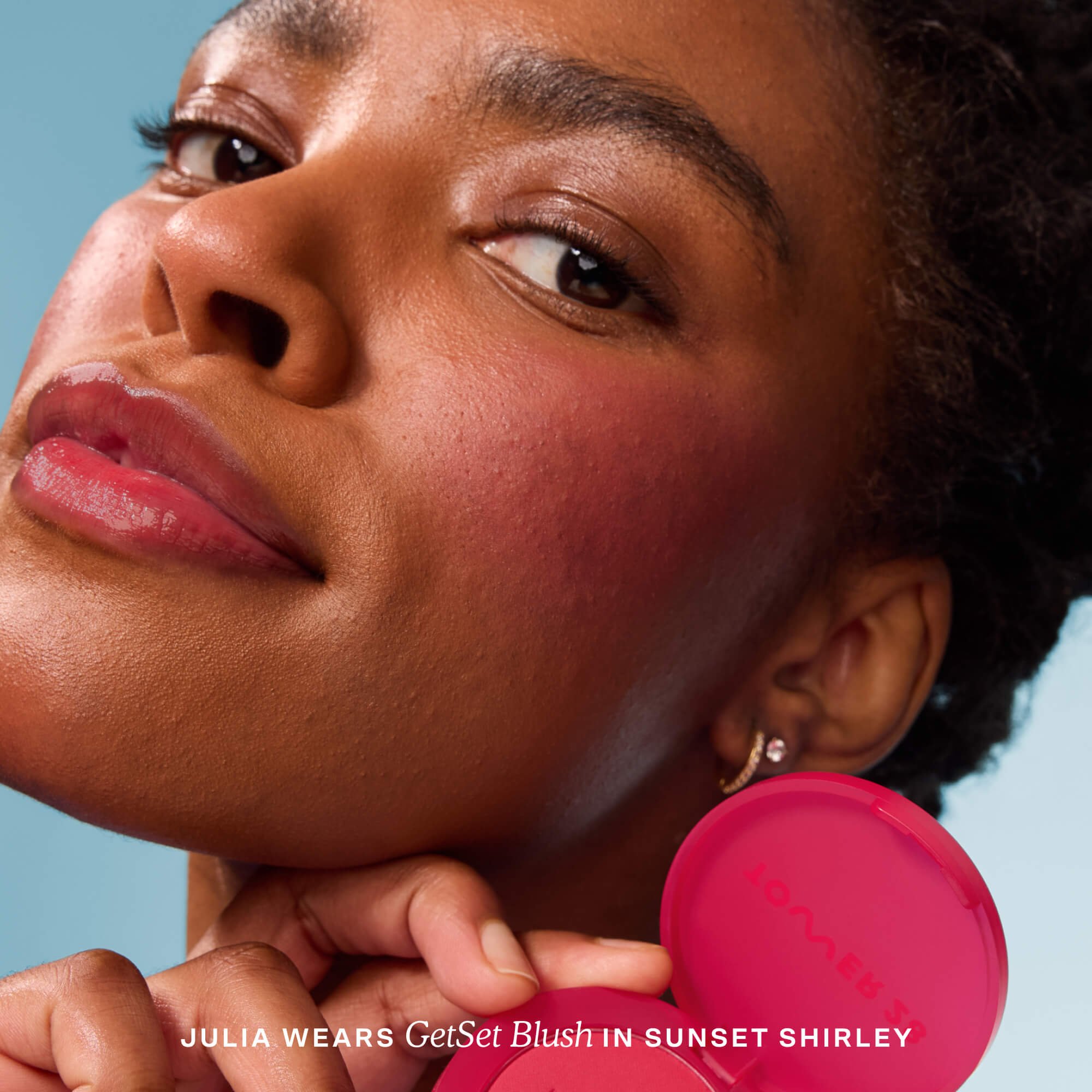Woman with pink blush on her cheek, holding a pink compact mirror against a blue sky.  [Sunset Shirley x Toni Bravo]