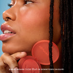 Close-up of a woman wearing red circular earrings with 'Tower 28' branding against a blue sky. [Downtown Daiquiri x Toni Bravo]